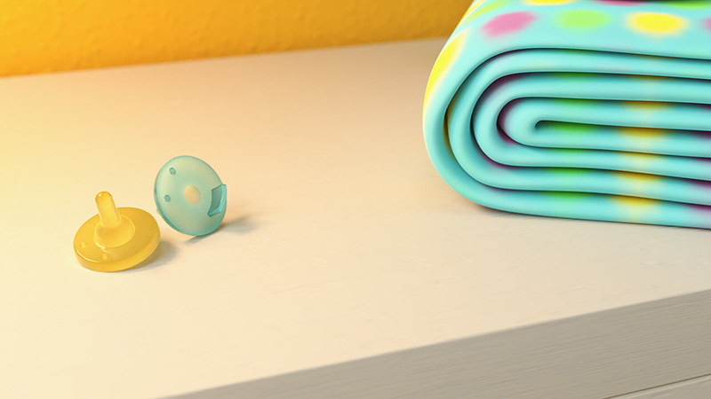 There are two one-piece pacifiers to the left of a blanket. All items are sitting on top of a chest of drawers.