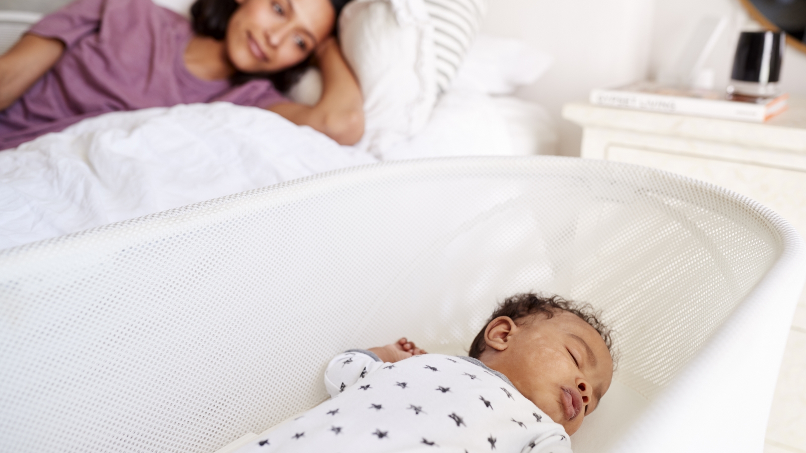 A mother is watching from her bed as a baby sleeps in a bassinet next to her.