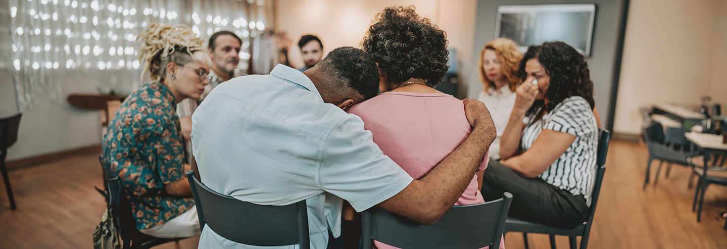 Two people sit with their arms around each other, while others sit in a circle around them expressing sadness.