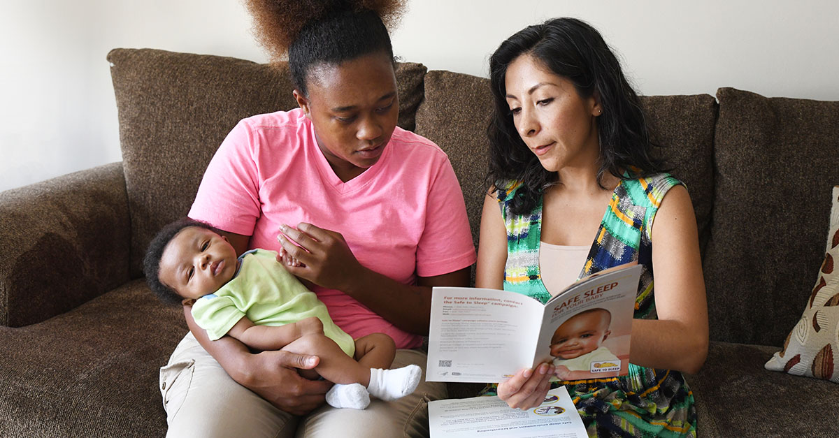 A mother holding a baby sitting on a couch talking with a community service professional. The community service professional is holding the Safe Sleep for Your Baby brochure.