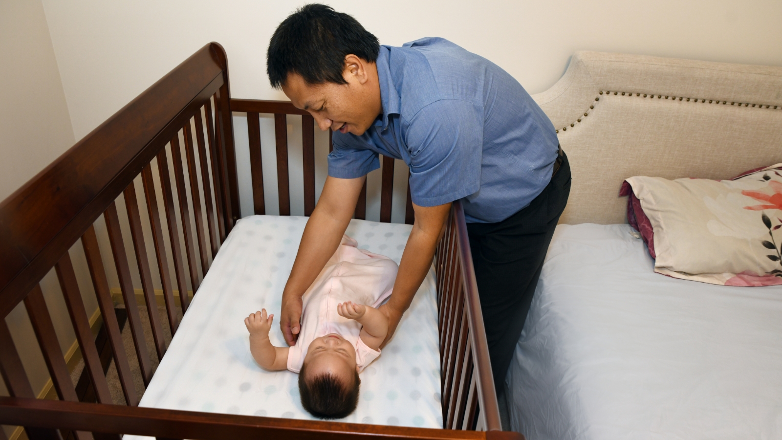 A father reaches toward a baby in a crib next to the bed.