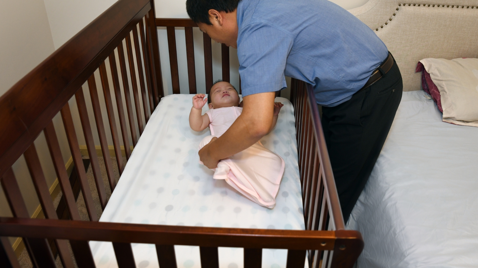 A father places a baby on their back to sleep in a crib next to an adult bed; the crib mattress is covered by a fitted sheet, and there are no other items in baby’s sleep area.