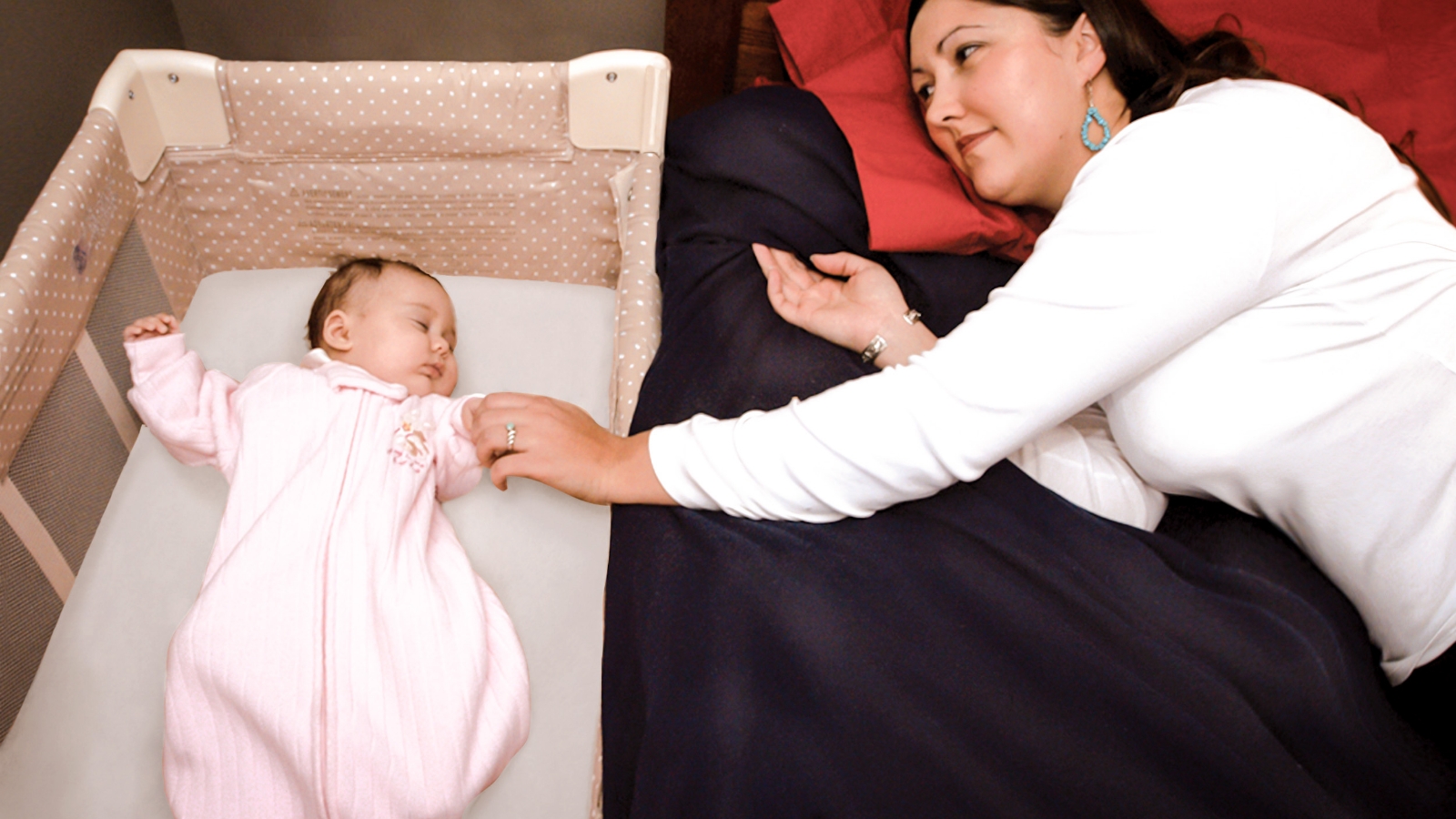 A mother and baby room sharing. Baby is sleeping on their back in their own sleep space, in the same room as the parents, next to the adult bed. The mother reaches toward the baby, who is close enough to comfort but not in the bed.