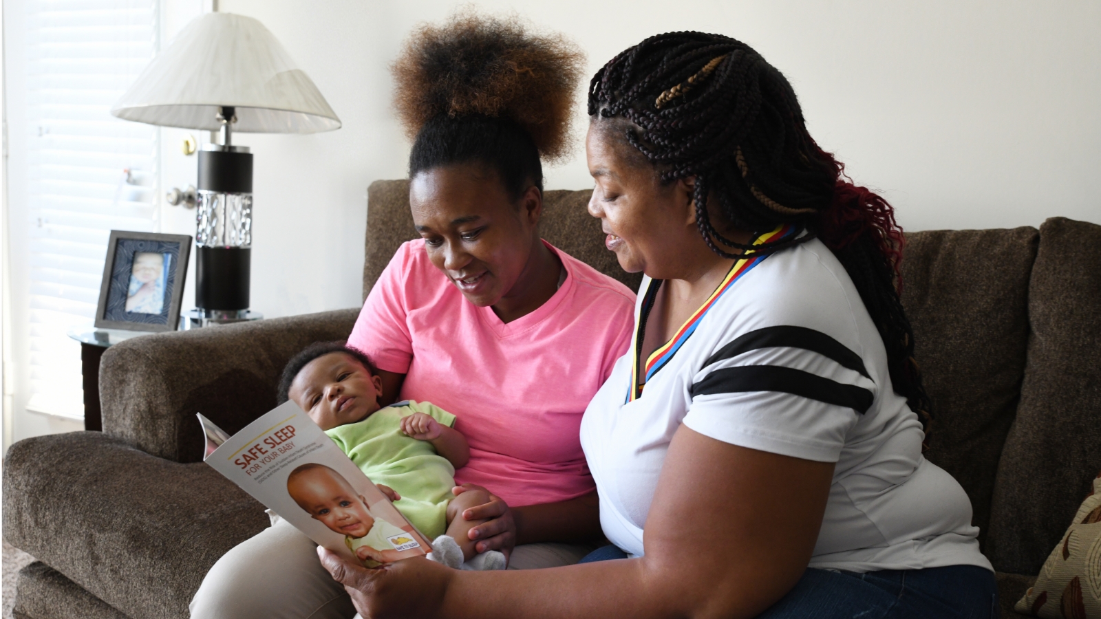 A mother (left) is holding a baby in her lap as another woman (right) goes over materials