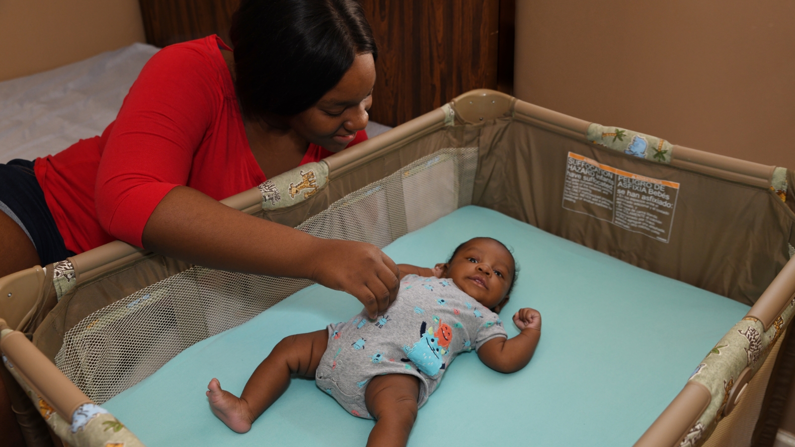 A woman reaches toward her baby, who is lying on their back in a crib next to her bed.