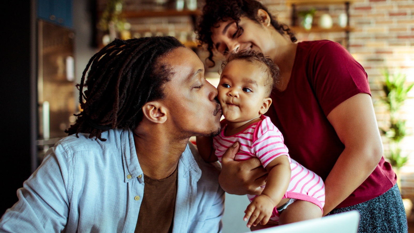 A caregiver (left) kisses a baby on the check while an additional caregiver (right) holds the baby in their arms.