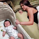 A baby sleeps on her back in a bassinet, which is next to her mother's bed. The mother lies in her own bed and rests her hand on the side of the baby's bassinet.