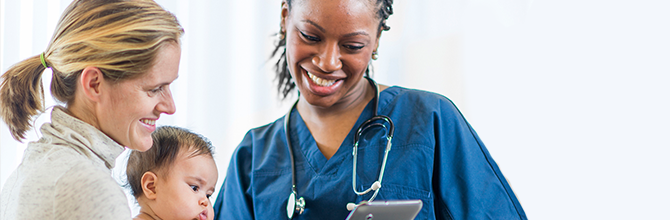 A health care provider shows a computer tablet to a mother who is holding her baby in her arms.