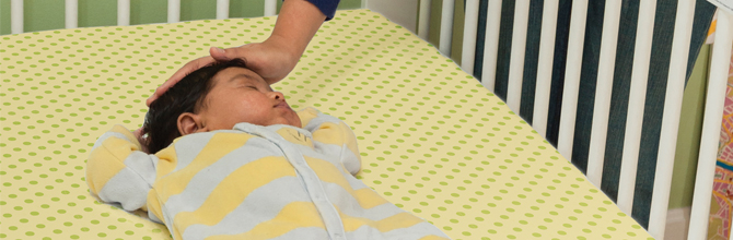 A baby sleeps on her back in a crib while her mother stands next to the crib and places her hand on the baby's head.