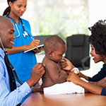 A baby focuses on their parent, who is distracting the baby’s attention while a healthcare provider examines them. Another healthcare provider stands nearby with a notepad.