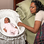 A baby sleeps on her back in a bassinet, which is next to her mother's bed. Her mother lies in her own bed and rests her hand on the side of the baby's bassinet.