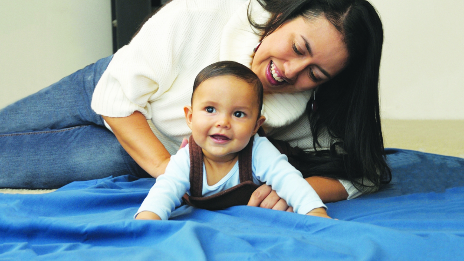 A baby lies on their stomach as their mother smiles and holds them from behind.